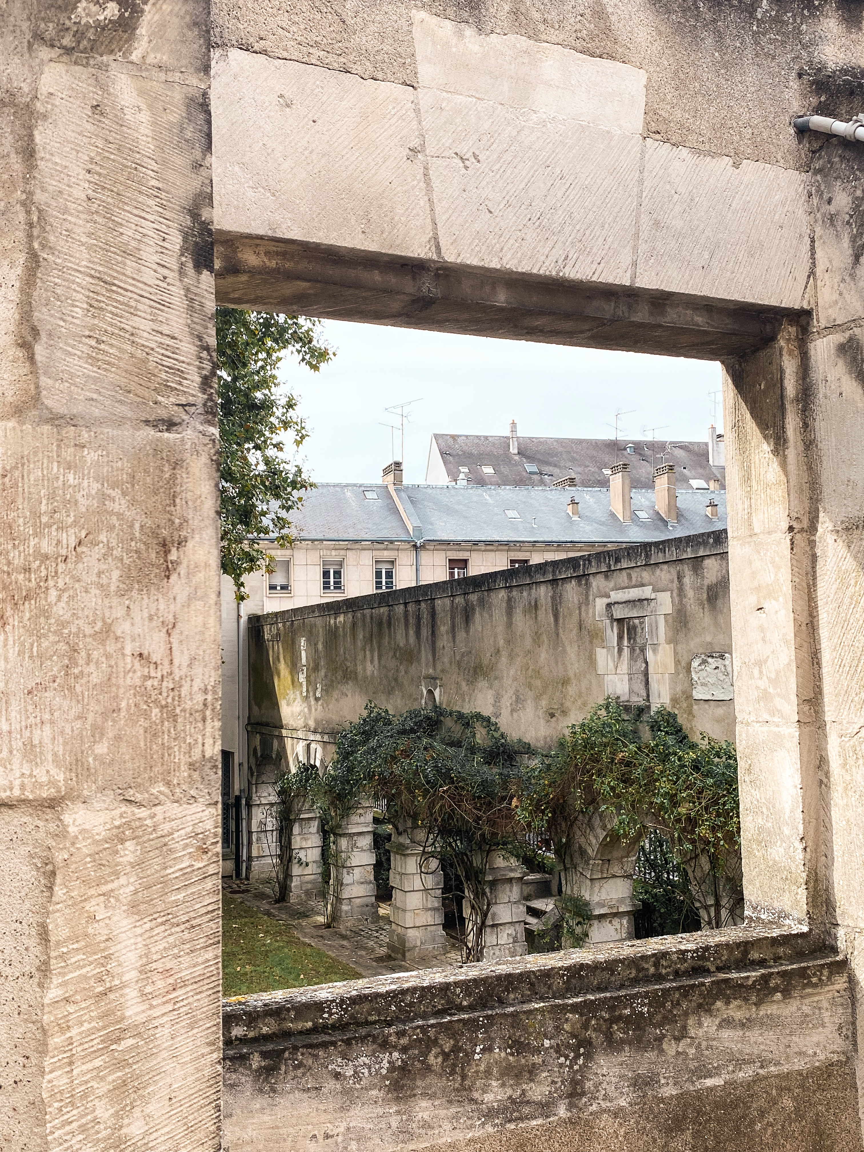 Encadrement de fenêtre en pierre pris en biais ; à l'intérieur du cadre un des côtés d'un cloître, et au fond des toits en ardoise