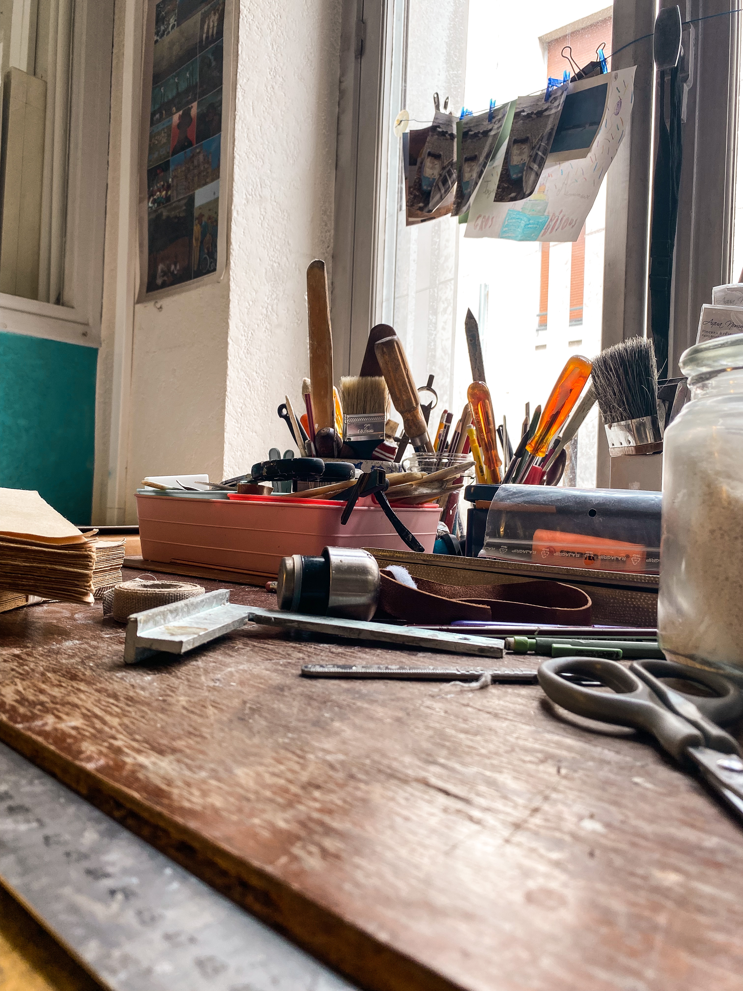 Atelier de reliure : Un bureau en bois sur lequel sont éparpillés différents types d'outils ; au fond, deux pots contenant l'un des pinceaux, l'autre crayons et tournevis. Derrière on voit une fenêtre avec des images accrochées sur un fil.