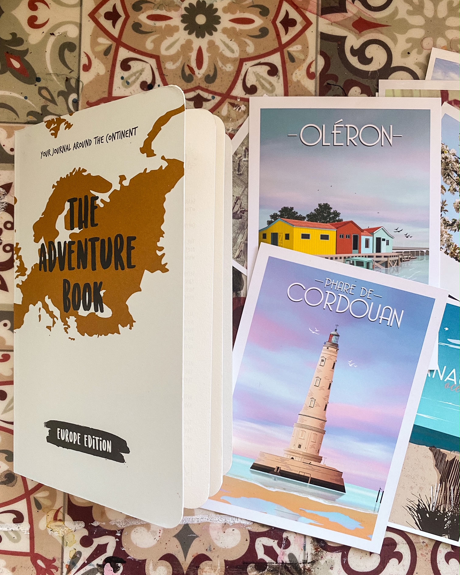 Photo de la couverture du livre The adventure book sur un fond imitant les carreaux de ciment. A côté deux cartes postales dessinées, l'une représentant le phare de Cordouan et l'autre des cabanes de pêcheurs colorées à l'île d'Oléron.