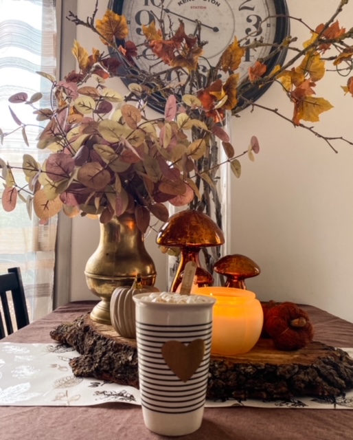 Un centre de table fait d'un rondin de bois sur lequel sont posés un vase doré contenant des feuilles d'eucalyptus, des faux champignons et des fausses citrouilles ; une bougie en forme de citrouille ; à l'arrière un vase de branches mortes avec une fausse guirlande de feuilles d'automne