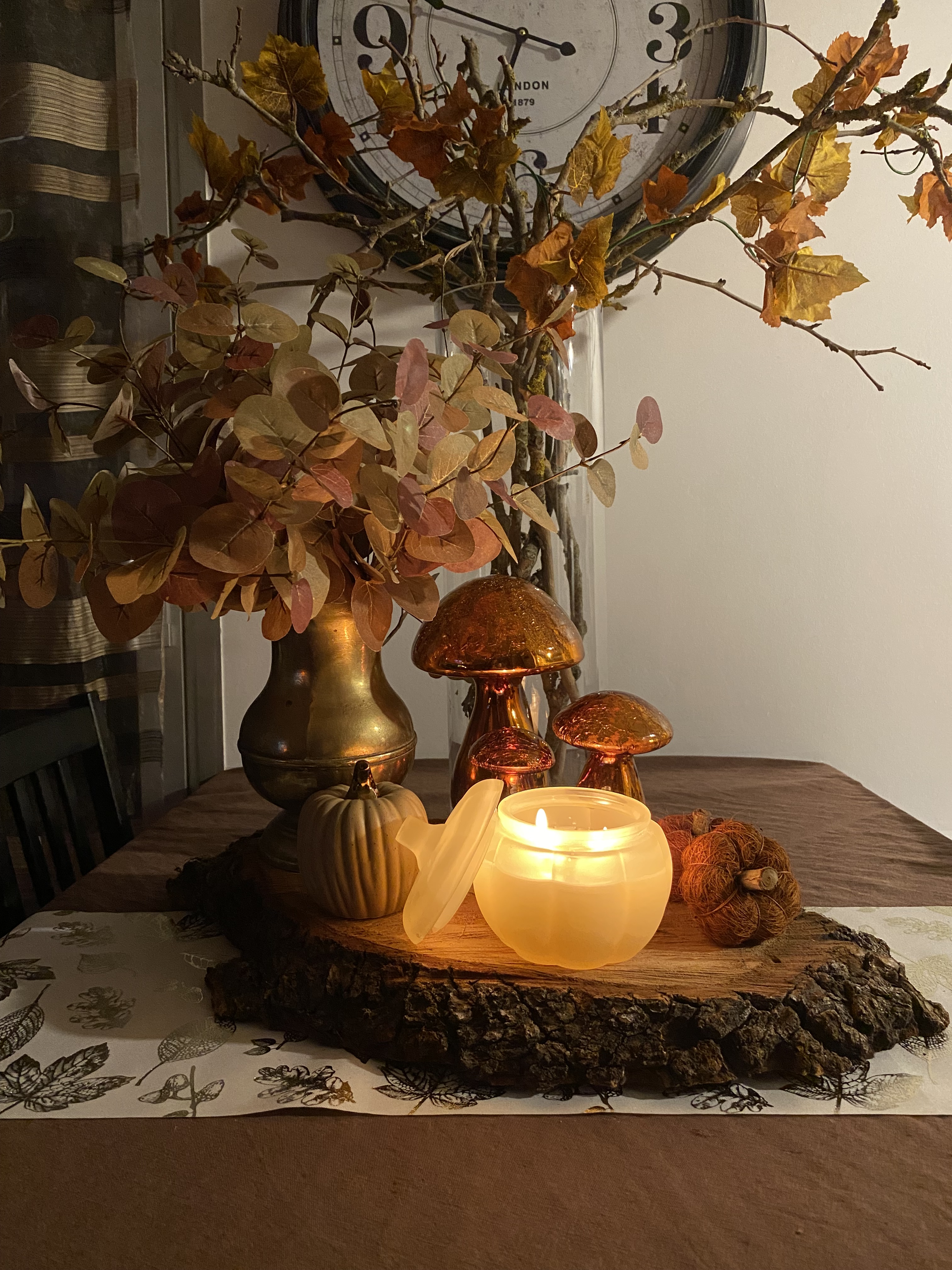 Un centre de table fait d'un rondin de bois sur lequel sont posés un vase doré contenant des feuilles d'eucalyptus, des faux champignons et des fausses citrouilles ; une bougie en forme de citrouille ; à l'arrière un vase de branches mortes avec une fausse guirlande de feuilles d'automne