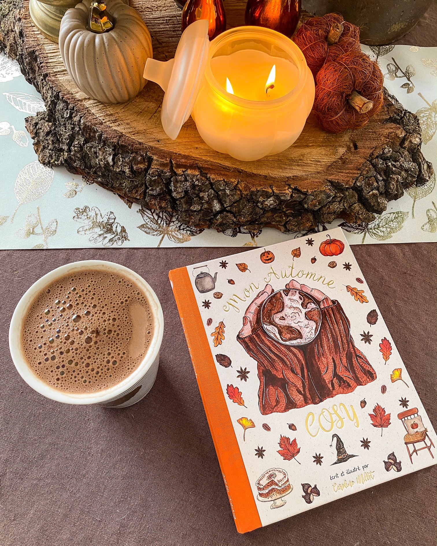 Le livre Mon automne cosy de Caroline Millet sur une nape chocolat ; à côté un mug de chocolat chaud ; en haut un centre de table fait d'un rondin de bois sur lequel est posée une bougie allumée en forme de citrouille et des petites citrouilles artificielles