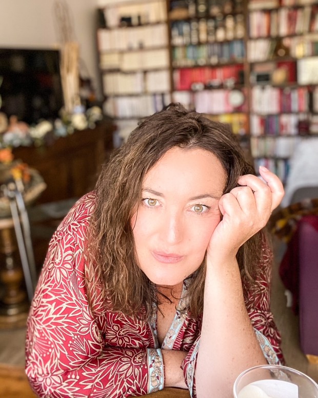 Portrait plan américain d'une femme châtain ; ses bras sont appuyés sur une table et sa maingauche est près de son visage ; une bibliothèque dans le fond